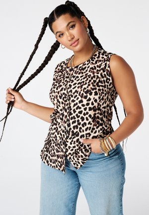 Vrouw met lange vlechten draagt een mouwloze top met luipaardprint, lichtblauwe jeans, gouden armbanden en oorringen, poserend tegen een witte achtergrond.