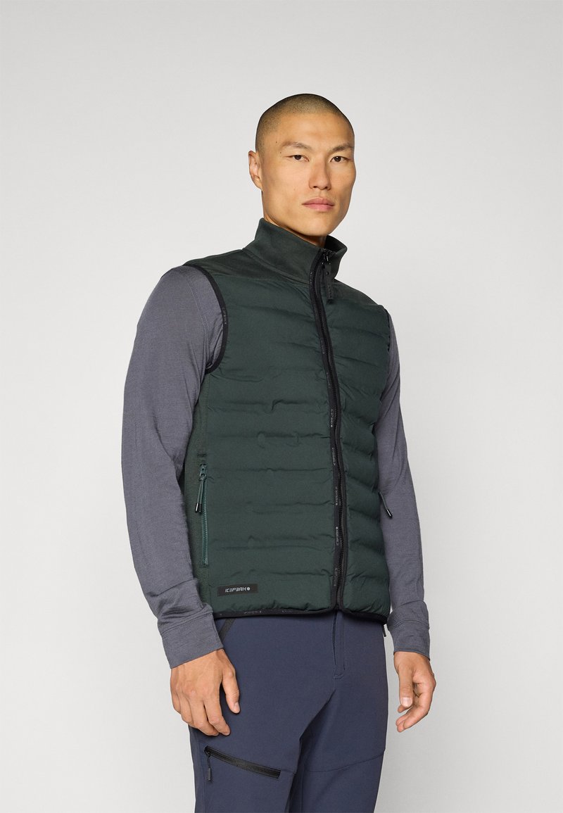 Gilet verde puffoso con colletto alto, chiusura a zip, tasche laterali e una texture liscia, indossato sopra una maglietta grigia a maniche lunghe.