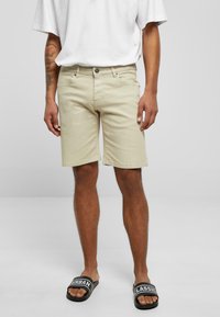 Hombre vestido con pantalones cortos beige hasta la rodilla, camiseta blanca y sandalias negras de tipo deslizar con el texto "URBAN" y "CLASSIC", de pie frente a un fondo sencillo.