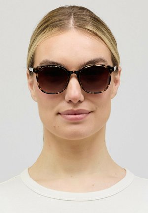 Vrouw met blond haar die ronde zonnebrillen met schildpadmotief en een wit shirt met ronde hals draagt, kijkt recht vooruit tegen een effen achtergrond.