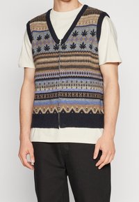 Pletená vesta s výstřihem do V, zdobená barevnými vzory v hnědé, modré a šedé, s předním zipem a tmavě námořnickým lemováním. Nasažená přes krémovou košili.