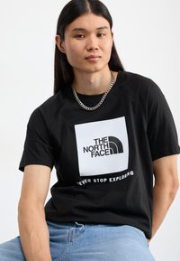 Camiseta de algodón negra con un logo cuadrado blanco que dice "THE NORTH FACE" y "NEVER STOP EXPLORING" en letras negritas. Manga corta, corte relajado.