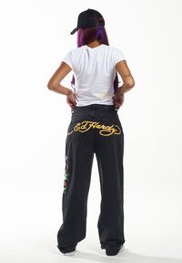 Mustat farkut, joissa on löysä malli, ja takana keltainen "Ed Hardy" -logo sekä brodeeratut kukkasuunnitelmat vasemmassa lahkeessa. Yllä valkoinen t-paita.
