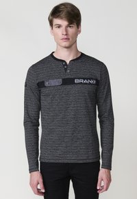 Majica dugih rukava crno-sive pruge s henley izrezom, s crnom horizontalnom trakom i detaljem logotipa preko grudnog dijela.