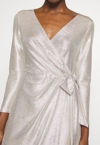 Metallisk silverfärgad omslagsklänning med långa ärmar, V-ringning och en knuten detalj vid midjan, med en texturerad, glittrande tyg.