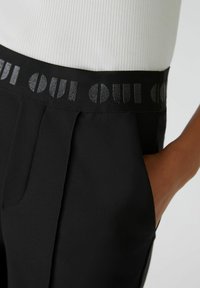 Zwarte broek met een gestructureerde tailleband met "OUI" in reflecterende letters, plooien aan de voorkant en zijzakken.