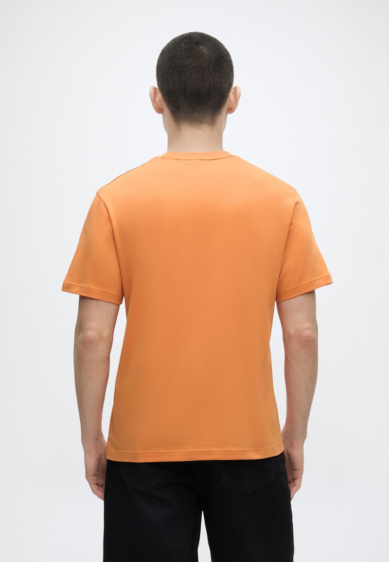 Man met kort haar, gekleed in een oranje T-shirt met korte mouwen en een zwarte broek, staat met zijn rug naar een effen lichte achtergrond.