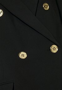 Gros plan sur un tissu noir croisé avec quatre boutons dorés brillants, deux boutonnières visibles et un détail de col plié.