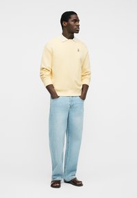 Hombre de pie con las manos en los bolsillos, vistiendo una camisa polo de manga larga color amarillo pálido, jeans azul claro y sandalias oscuras sobre un fondo liso.