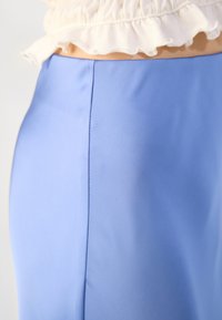 Gros plan d'une jupe bleu clair avec une couture verticale, portée avec un haut blanc à volants laissant apparaître une petite partie du ventre.