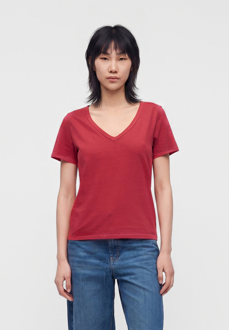 Femme debout face à l'avant, portant un t-shirt rouge à col en V et manches courtes ainsi qu'un jean bleu, sur un fond uni clair.