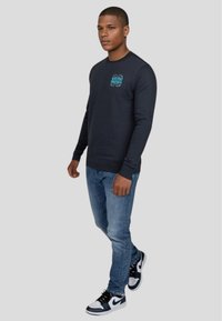 Mörk marinblå långärmad sweatshirt med en turkoss grafisk design, kombinerad med ljusblå jeans och svarta och vita sneakers.