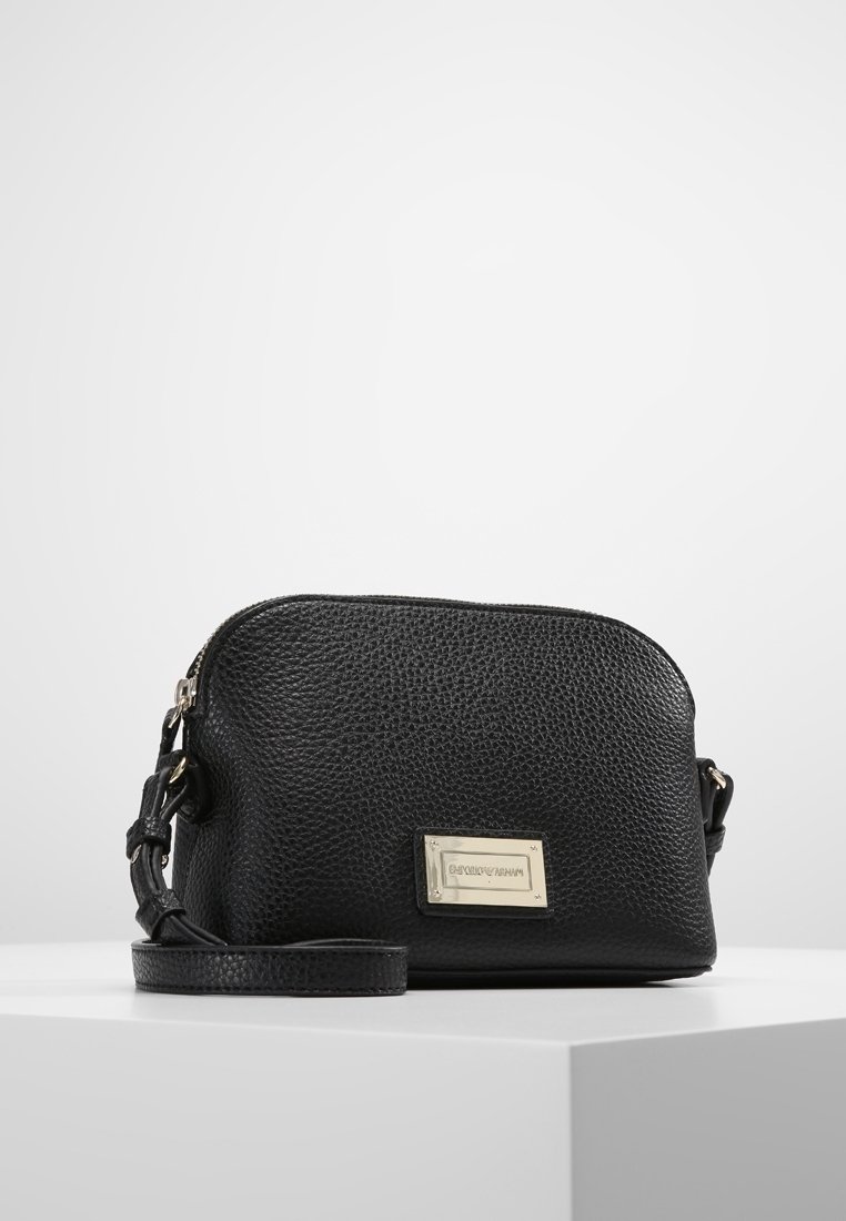 Černá kožená Crossbody taška s texturou, s nastavitelným popruhem a obdélníkovou stříbrnou kovovou značkovou plakietou na přední straně, vystavená na bílé podstavě.