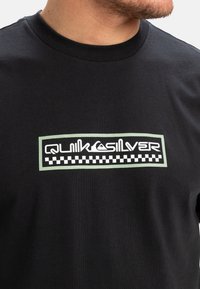 Czarny bawełniany t-shirt z białym i zielonym logo Quiksilver w prostokątnej ramce, z czarno-białą kratką na dolnej krawędzi.