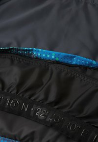 Schwarzes Kleidungsteil von innen, das einen leuchtend blauen Stoff mit einem Punktmuster zeigt. Verfügt über einen sichtbaren Reißverschluss und eingestempelte Koordinaten in Schwarz.
