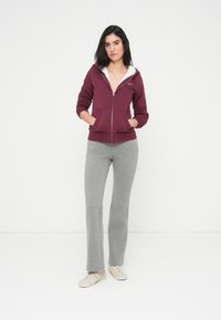 Μπορντό φούτερ με φερμουάρ και εσωτερική επένδυση φλις στην κουκούλα, δύο μπροστινές τσέπες, σε συνδυασμό με γκρι φουσκωτό παντελόνι φόρμας και ανοιχτό μπεζ αθλητικά παπούτσια.