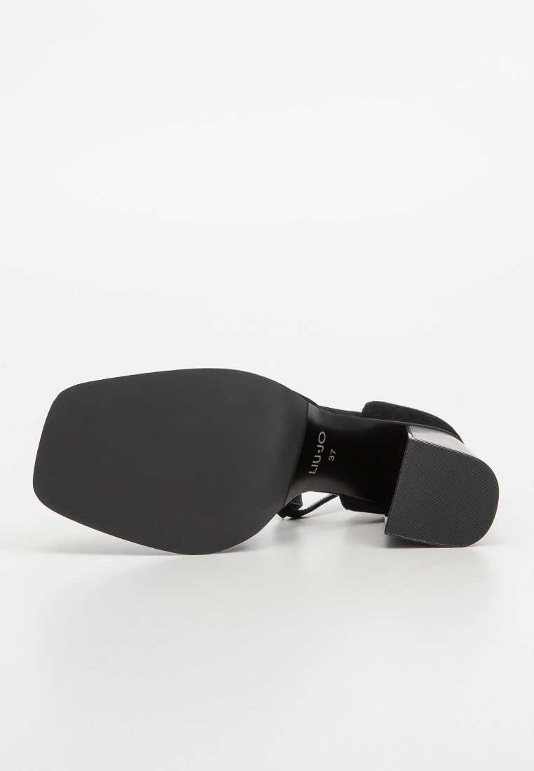 Zapato de tacón alto negro con suela ancha y plana, parte superior de tela, textura mate y un acento brillante en el tacón. Detalles de tiras alrededor del tobillo.
