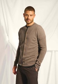 Hnědý kardigan vyrobený z měkkého materiálu s kulatým výstřihem, zapínáním na knoflíky a pružnými manžetami. Nosený přes černé tričko.