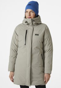 Lichtgrijze parka met een hoge kraag, ritszakken en een zwart logo. Gecombineerd met een blauwe beanie. Heeft een pufferontwerp en eenvoudige lijnen.