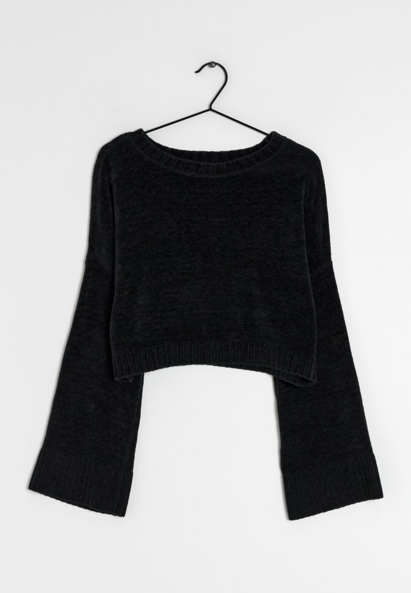 Pull court noir à manches longues avec larges poignets et ourlet côtelés, suspendu sur un cintre en fil noir sur un fond blanc.