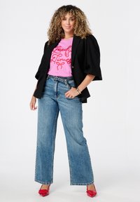Femme aux cheveux bouclés portant un blazer noir, un t-shirt rose "Good Times", un pantalon large et des talons rouges, se tenant avec une main sur la hanche.