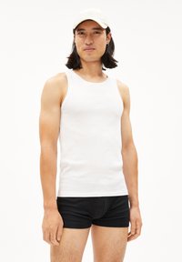 Witte katoenen tanktop met brede armsgaten, gedragen met zwarte strakke shorts. Model heeft lang donker haar en draagt een beige pet.