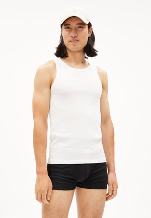 Weißes Baumwoll-Top mit weiten Armausschnitten, kombiniert mit schwarzen, eng anliegenden Shorts. Das Model hat lange, dunkle Haare und trägt eine beige Cap.
