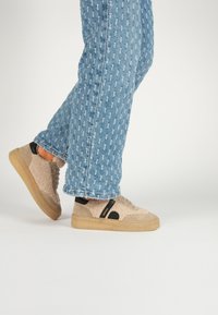 Beige textured sneakers with black accents and a gum sole, paired with blue denim jeans featuring a small repeated pattern.