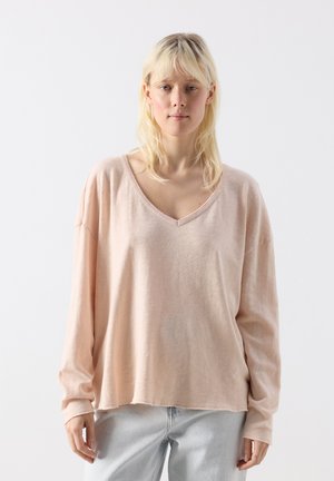 Jauna blondinė moteris dėvi laisvą, šviesiai rožinę V formos ilgomis rankovėmis palaidinę ir šviesiai mėlynas džinsus, stovinti prieš baltą foną.