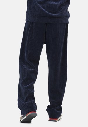 Persona che indossa pantaloni di corda blu navy larghi e scarpe da ginnastica casual multicolori, in piedi contro uno sfondo semplice.