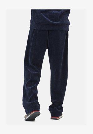 Persona che indossa pantaloni di corda blu navy larghi e scarpe da ginnastica casual multicolori, in piedi contro uno sfondo semplice.