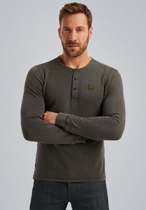 Chemise henley à manches longues de couleur vert olive avec un col rond, trois boutons et un petit logo carré brodé sur la poitrine.
