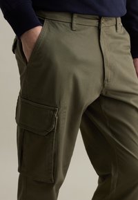 Olivegrüne Cargo-Hosen aus Baumwollmischung, mit einer Ziehentasche und seitlichen Patches, gerader Schnitt und glatter Textur.