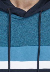 Huppari, jossa on teksturoitu teal-väri ylhäällä, tasainen sininen keskiosa ja valkoisia ja laivastonsinisiä vaakaraitoja alaosassa. Nyöri yksityiskohtana.