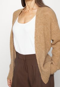 Cárdigan de punto beige con mangas largas, frente abierto y textura suave, usado sobre una camiseta sin mangas blanca y pantalones marrones.