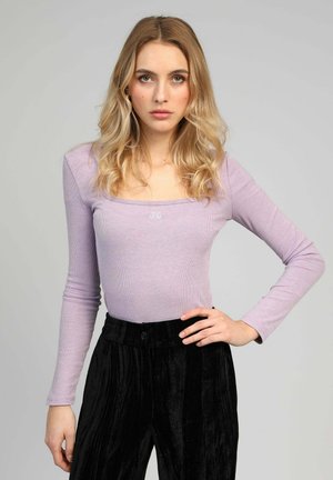 Femme blonde aux cheveux ondulés portant un haut lilas à manches longues et un pantalon en velours noir taille haute, debout avec une main sur la hanche.