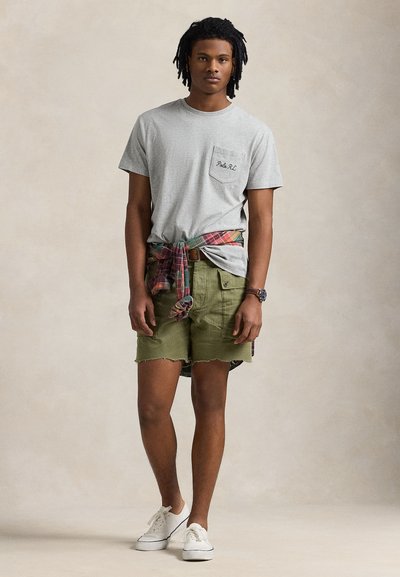 Young man standing, wearing light grey t-shirt, green cargo shorts, plaid shirt tied around waist, white sneakers, and a wristwatch.