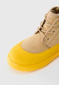 Bottine en daim beige avec bout et semelle en caoutchouc jaune, présentant des lacets ronds beiges et des œillets en métal, sur un fond clair.