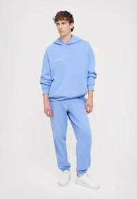 Lyseblå hoodie og sweatpants sæt lavet af blødt materiale. Hoodien har snøre og et subtilt logo. Modellen har hvide sneakers på.