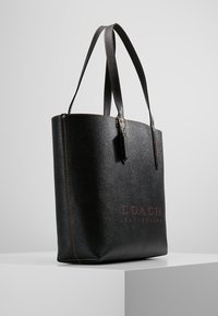 Czarna skórzana torba tote z teksturowanym wykończeniem, z wyróżniającym się logo, przeszywanym konturem i długimi uchwytami.