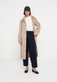 Long manteau trench beige avec fermeture à boutons, porté sur un haut blanc à motifs et un pantalon bleu marine ; une casquette noire et des chaussures plates complètent le look.