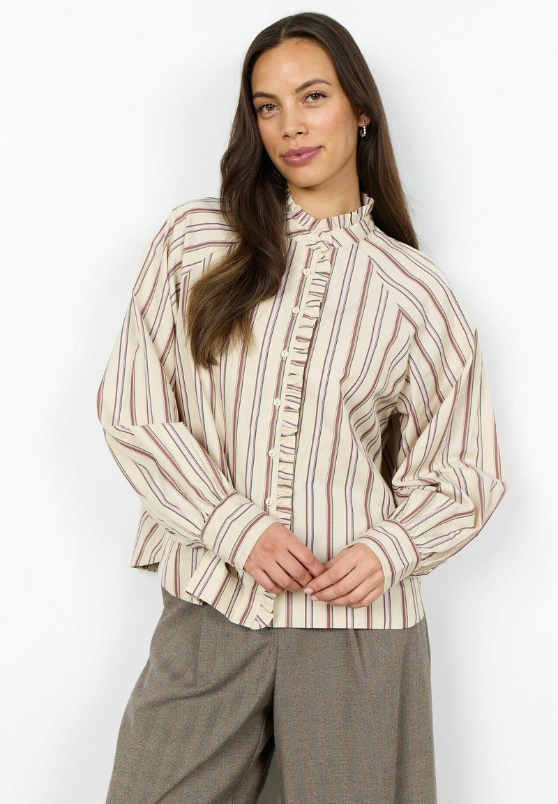 Femme portant un chemisier crème à rayures verticales rouges et noires avec un détail volanté, associé à un pantalon gris ample, debout sur un fond blanc.