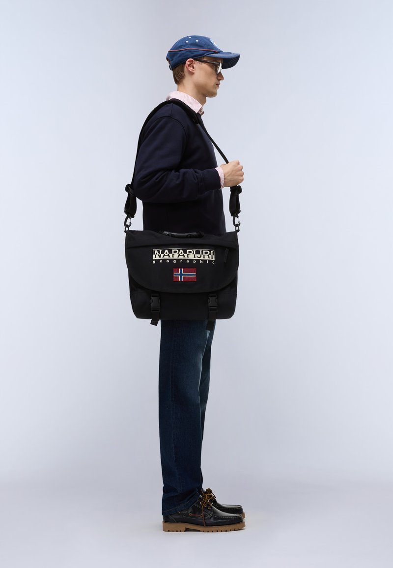 Homme portant des lunettes de soleil, une casquette bleue, un sweat-shirt bleu marine, un jean et des chaussures marron, portant un sac messager Napapijri Geographic noir.