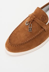 Zapato estilo mocasín de ante marrón con punta redondeada, que presenta un encanto de logo plateado y costuras decorativas a lo largo del borde.