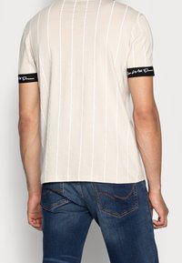 Béžové pruhované tričko s krátkými černými rukávy, které obsahuje bílé pruhy a text. Bavlněný materiál, slim fit, minimalistický design.