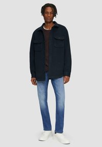 Marineblaue Jacke mit Knöpfen und zwei Brusttaschen, über einem dunklen Pullover getragen, kombiniert mit hellblauen Jeans und weißen Sneakers.