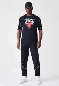Črn športni komplet, ki vključuje majico s kratkimi rokavi z rdečim logotipom Chicago Bulls in črne trenirke z belimi detajli ter logotipom.