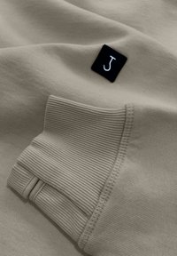 Beige sweatshirt fabric with ribbed cuffs. Black tag with a white 'J' logo. Soft texture and smooth finish.