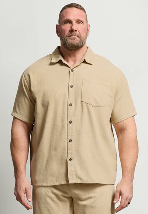 Bärtiger Mann, der ein beiges, kurzärmeliges Hemd mit Knöpfen und passende Hosen trägt, steht vor einem einfarbigen hellen Hintergrund.