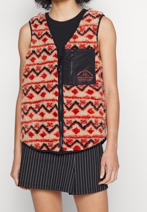 Ærmeløs, fluffy vest med zigzag-mønster i rød, sort og beige, frontlynlås og sort brystlomme med lynlås og orange tekst, båret over en sort top.